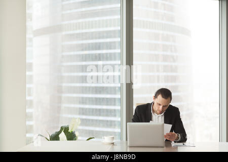 Company leader examines document at desk in modern office with large windows. Businessman reading important notice at workplace. Entrepreneur focused Stock Photo