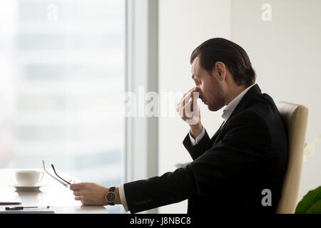 Tired businessman at desk holding eyeglasses in hand and massaging nose bridge. Man feels fatigue in eyes after hard work, discomfort because of long Stock Photo