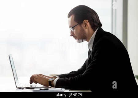 Side view portrait of stylish businessman in glasses typing on laptop. Successful entrepreneur working on company marketing strategy, analyzes new mar Stock Photo