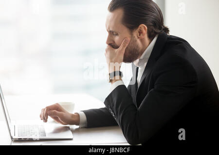 Businessman yawning while working on computer at workplace. Male entrepreneur suffering from lack of sleep, chronic fatigue and drowsiness at desk wit Stock Photo