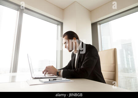 Serious businessman working on laptop at desk in office. Focused young entrepreneur works on computer of an important project. Man carefully examines Stock Photo