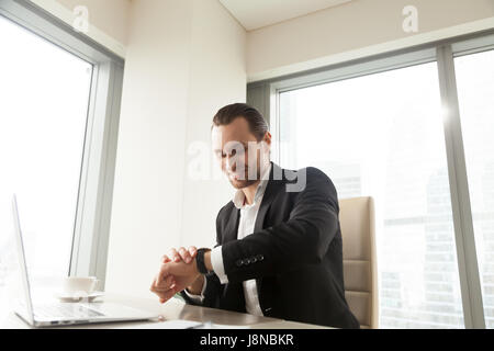 Successful businessman looking at wristwatch while sitting at desk with laptop in office. Company leader checks time remaining to start online confere Stock Photo