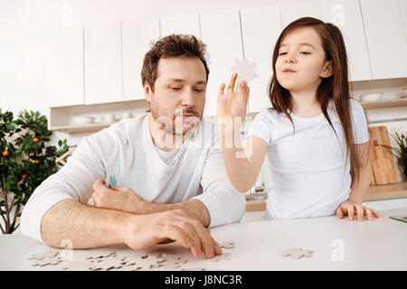 Quality time. Single young father doing puzzle with his little daughter holding a jigsaw piece in one hand and watching on it while he trying to arran Stock Photo