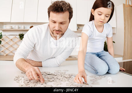Captivated by puzzles. Delicate pretty girl sitting on her knees on the kitchen counter and doing a jigsaw puzzle together with her father, both being Stock Photo