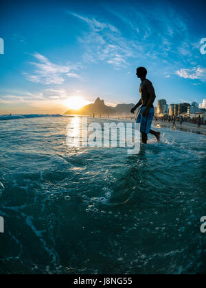 Young Brazilian man walking in silhouette through the waves on Ipanema Beach as the sun sets behind Two Brothers Mountain in Rio de Janeiro, Brazil