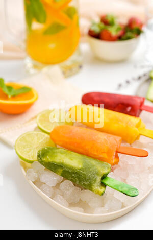 closeup of some different homemade natural ice pops served in a plate with ice, placed on a table with a jar of juice and a bowl with strawberries Stock Photo