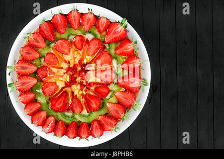 Fresh fruit pie decorated with strawberries, peaches and kiwi, on black wooden background, top view shot with copy space. Stock Photo