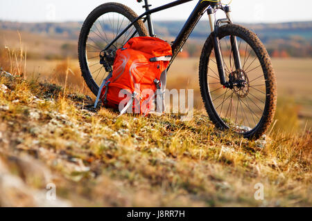 Mountain bike after ride in nature with backpack stands on the hill. Sunny day in the countryside. A bicycle without people on a background of plane. Stock Photo