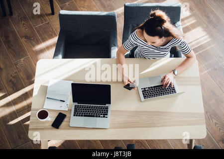 Busy young woman texting with mobile phone and writing notes while sitting at her desk. Pretty caucasian female working in home office or a cafe with her laptop. Stock Photo