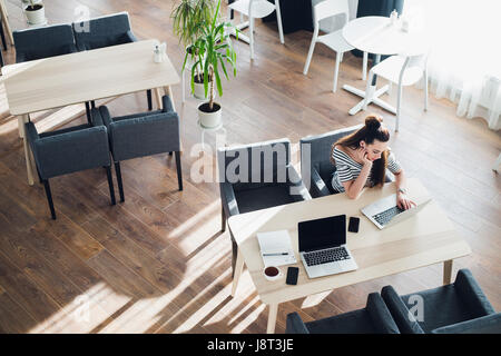 Young woman casually working on her laptop in a cafe. Bored attractive female sitting with a cup of coffee at work. Stock Photo