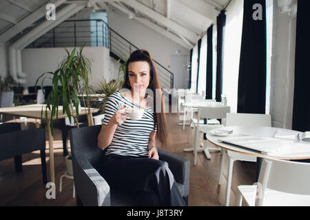 Beautiful young lady holding a cup of coffee looking pensive and absorbed in her thoughts, leaning on her elbows while sitting in a sidewalk cafe. Stock Photo