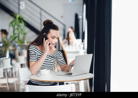 Beautiful adult woman in striped shirt is talking on the phone, holding a cup and smiling while working with a laptop in a cafe. Stock Photo