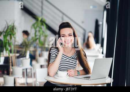 Beautiful adult woman with beautiful smile is talking on the phone and working with a laptop in a cafe while looking away. Stock Photo