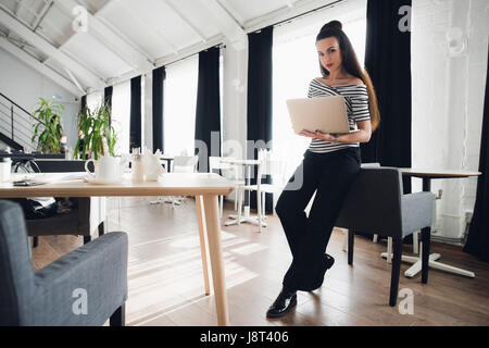 Picture of a beautiful female using laptop while stending in a cafe near table. Young businesswoman sitting in a lounge and working on a personal computer. Stock Photo