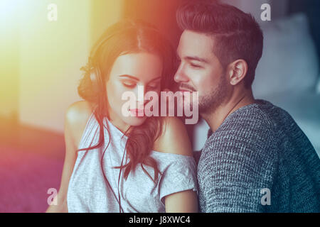 Young couple listening music by headphones together at home in sunset, sharing music Stock Photo