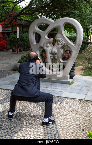 Guilin, China.  Elephant Trunk Hill Park.  Man Photographing Two Friends behind Modern Sculpture Representing Love. Stock Photo