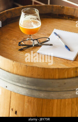 Glass of wheat beer with a pair of glasses, pen and notepad standing on a wooden barrel in a brewery Stock Photo