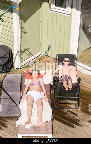 Female teenager and young boy tanning on sun loungers outdoors with water sprinkler sprinkling from above  Model Release: Yes.  Property release: No. Stock Photo