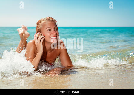 Beautiful young woman enjoying on the beach and listening to sea shells. She is smiling and pensive looking away. Stock Photo