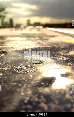 3d rendering of raindrops on street puddle in front of blurred sunshine and city Stock Photo