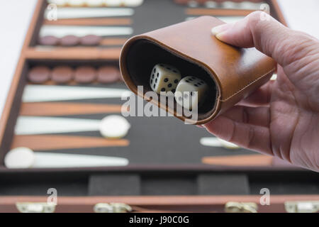 Readying to roll backgammon dice on the brown and cream backgammon board blurred in the background. Focusing on the dice and dice cup in hand. Stock Photo