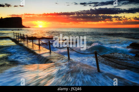 Warm light of rising sun above horizon highlights fast waves of Pacific ocean flowing over the rock pool at Avalon beach of Sydney northern beaches. Stock Photo
