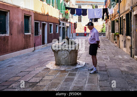 Venice,Castello. Narrow picturesque street with weathered houses,old water well, washing lines,laundry day and senior man with cat Stock Photo