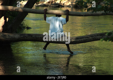 Boy alone sitting on fallen tree trunk over water Stock Photo