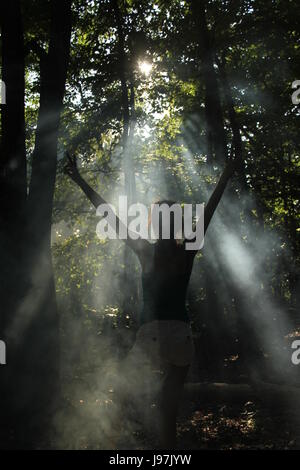 Young girl with arms raised facing sun beams coming through the trees in the woods Stock Photo