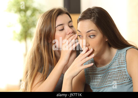 Upset girl because a friend is telling secrets to the ear sitting on a couch in the living room at home Stock Photo