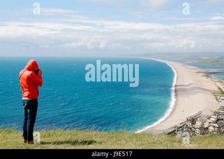 Young man in jeans and red jacket takes photos of Weymouth and coastline from a viewing platform on the Isle of Portland, Dorset, UK. Stock Photo