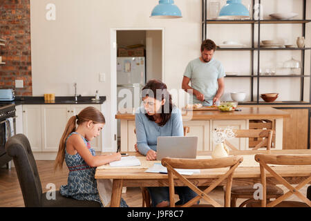 Mother Helps Daughter With Homework As Father Makes Meal Stock Photo