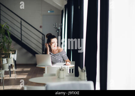 Looking for inspiration. Young beautiful pensive woman making some notes and looking through window while sitting in chair at her working place. Stock Photo