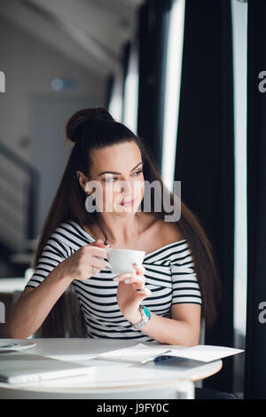 Young lady drinking coffee and looking through a window while sitting in a cafe. Stock Photo
