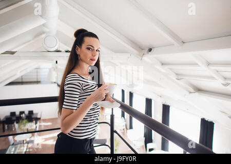Close up portrait of a girl with brown hair holding take away coffee cup and smartphone while looking at the camera. Stock Photo