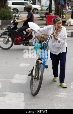 THAILAND, CHIANG MAI, NOV 07 2014, Old woman push a bicycle across a street in the city center. Stock Photo