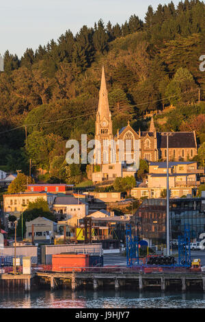 Port Chalmers, New Zealand - March 15, 2017: Iona Church under sunrise light towers over the harbor. Green forested hill in back. Houses, traffic and  Stock Photo