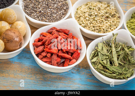 collection of superfoods in small ceramic bowls against rustic wood: macadamia nuts, hemp seed hearts, goji berry, stevia herb and chia seeds Stock Photo