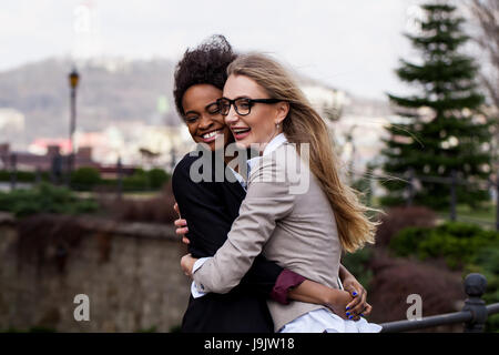 Two smiled happy businesswomen hugging each other in the park. Women wearing suits. One woman is black. Stock Photo