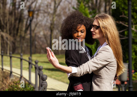 Two attractive and successful businesswomen doing selsie in the park. One woman is black.. Stock Photo