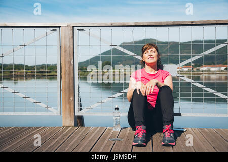 Middle aged woman relaxing after running enjoying the sun leaning on a fence Stock Photo