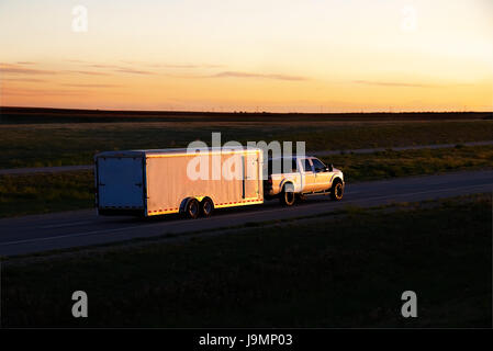 A white Pickup truck Pulls and unmarked box-trailer down a rural highway during late sunset. All visible markings and trademarks have been removed. Stock Photo