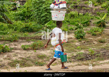 Woman with young child on back walks with huge bucket filled with clean pots and pans on her head Stock Photo