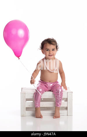 A happy, one year old, baby girl holding a pink balloon. She is wearing metallic pink leggings, a string of pearls and sitting on a wooden crate on a  Stock Photo