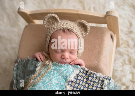 Two week old newborn baby boy wearing a tan, crocheted, bear bonnet. He is sleeping on a tiny, wooden bed and covered with a blue quilt. Stock Photo