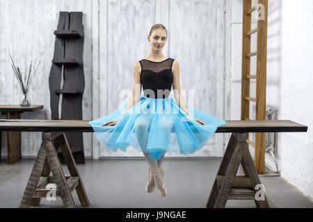 Smiling ballerina sits with crossed legs on the wooden table on a light wall background in a studio. She wears a black leotard with a cyan tutu and po Stock Photo