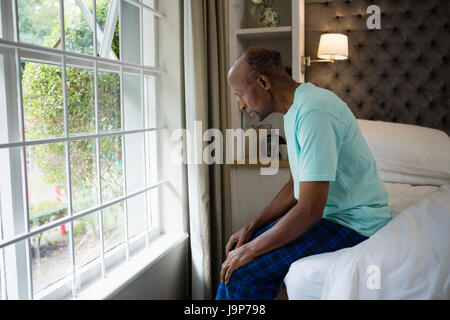 Side view of thoughtful senior man sitting on bed by window in bedroom at home Stock Photo