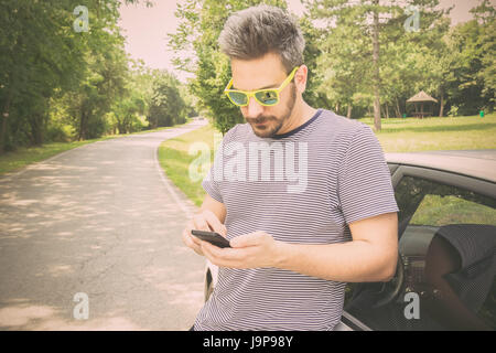 Tourist guy standing next to car and looking at smart phone. Driver using road map navigation app or calling road assistance service. Travel and techn Stock Photo