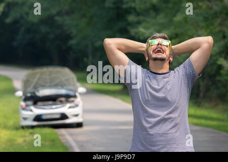 Stressed and frustrated driver pulling his hair while standing on the road next to broken car. Road trip problems and assistance concepts. Stock Photo