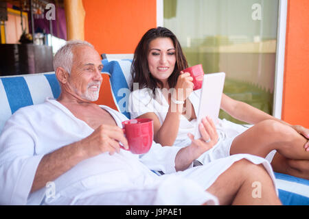 Older man and young woman using a tablet computer while enjoying coffee in bathrobes on loung chairs, Nuevo Vallarta, Nayarit, Mexico Stock Photo